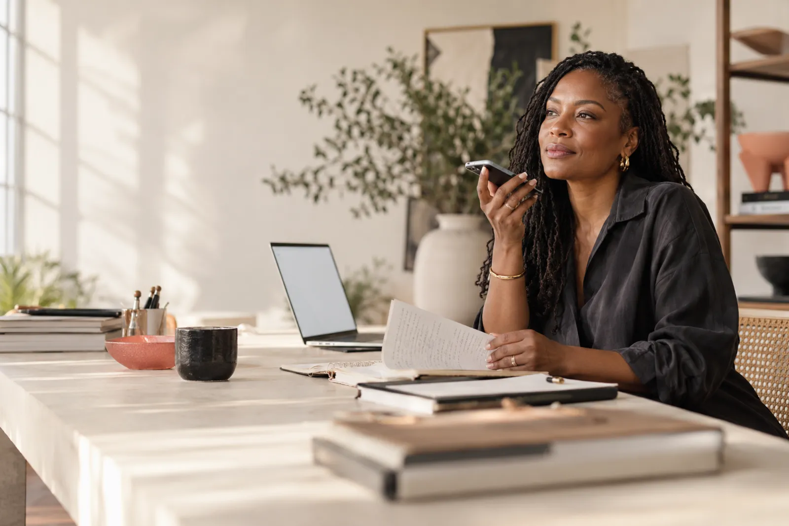 A business owner at her desk records an idea on her phone, with a notebook open in front of her.
